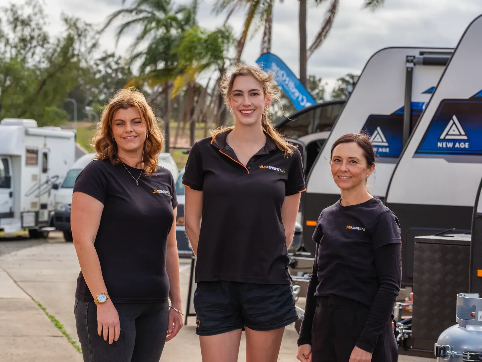 Three Kennedy RV team members in branded shirts standing in front of caravans on the yard