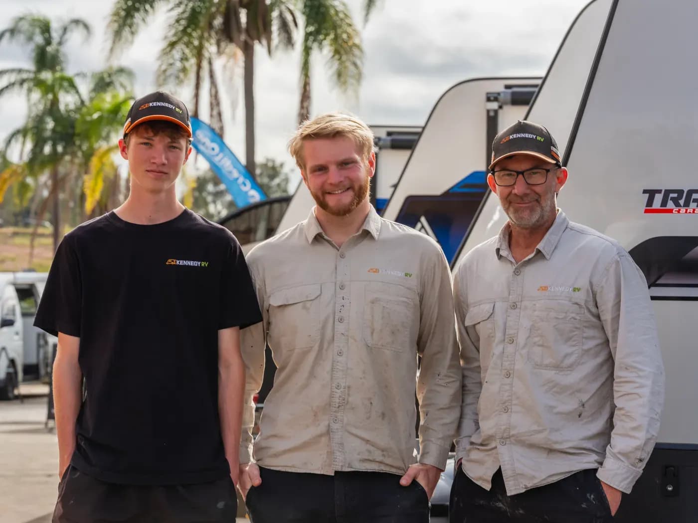 Three Kennedy RV team members in branded work shirts standing in front of a caravan on the yard